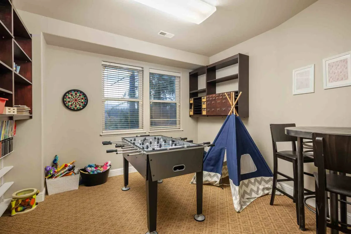 This image captures a vibrant game room featuring a foosball table, play tent, bookshelf, and a dartboard. The space is filled with natural light, and various entertainment options are visible, suggesting an area for both energetic play and quiet moments like reading.