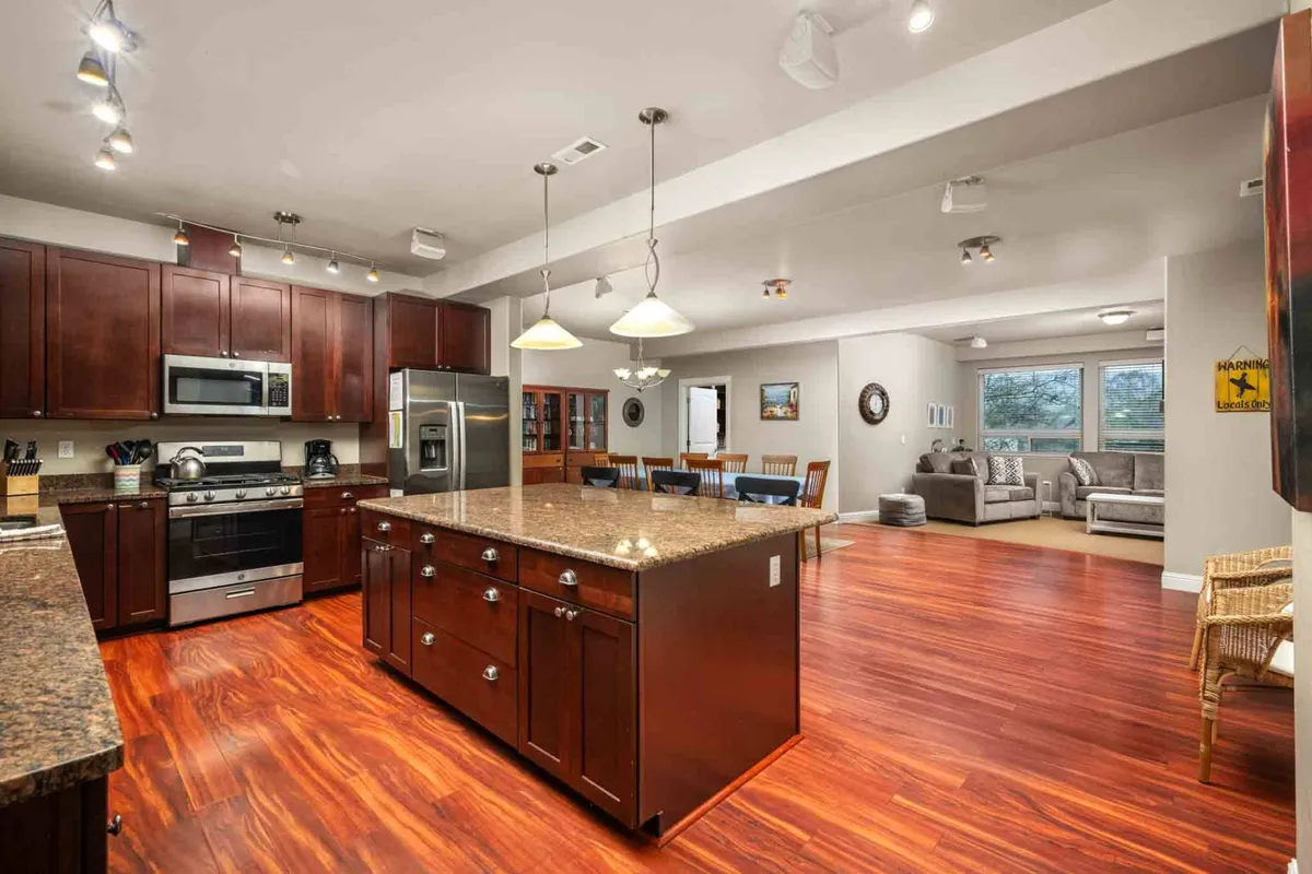 The image showcases a bright, open-concept kitchen and living space, featuring a gleaming granite island, stainless steel appliances, a warm wooden dining table, and an inviting living area with plush seating. Sunlight pours in, highlighting the thoughtful design that encourages gathering and relaxation.