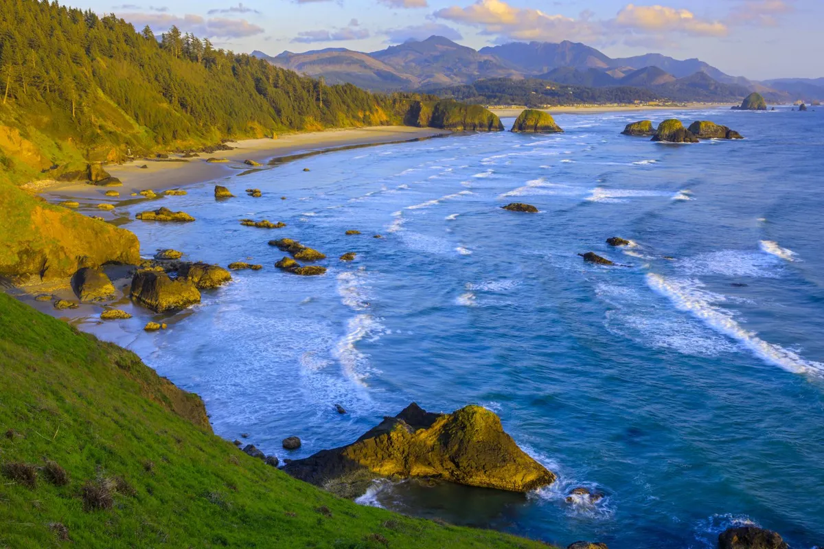 A breathtaking view of ocean waves rolling onto a sandy beach, with dramatic rock formations and forested mountains visible in the distance. The scene captures the expansive coastline, perfect for walks, beachcombing, or yoga in the fresh ocean air.