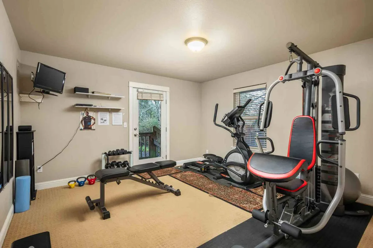 A fully equipped home gym featuring exercise machines, free weights, a television, and a doorway that brings in natural light. The space is designed for both energizing workouts and post-exercise relaxation, blending fitness with comfort.