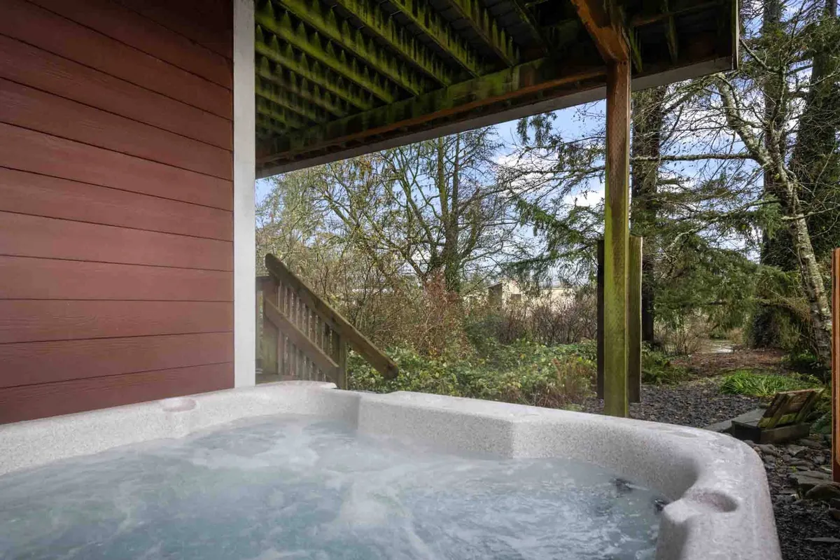 This exterior image features a bubbling hot tub situated on a private deck, surrounded by tall trees, with an outdoor staircase visible nearby. The setting is tranquil and immersed in nature, offering a glimpse of the relaxing and rejuvenating outdoor experience described in the section.