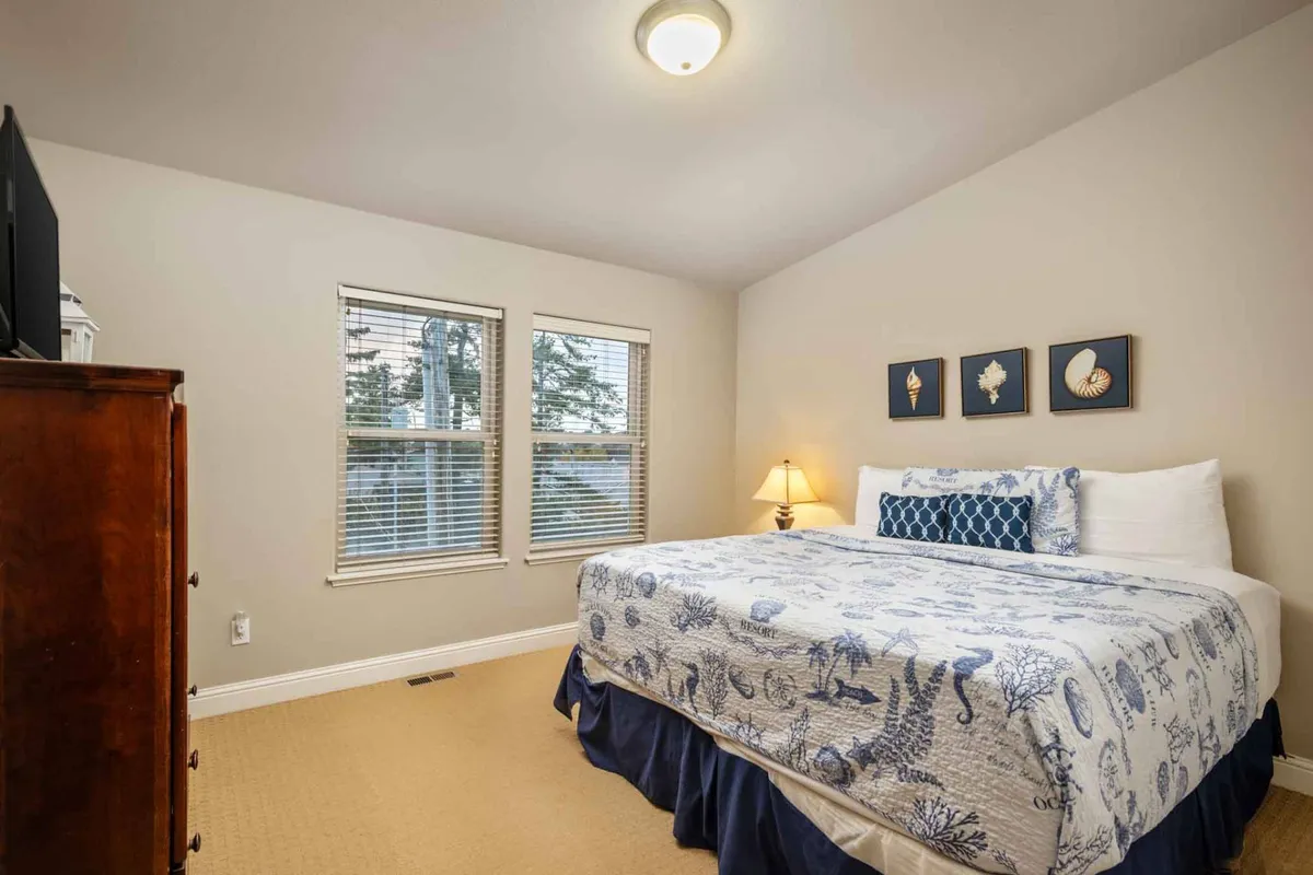 A serene bedroom featuring a bed with ocean-themed bedding, framed sea shell artwork above the headboard, and a wooden dresser. Soft natural light fills the room, creating a peaceful and restful atmosphere ideal for relaxation.