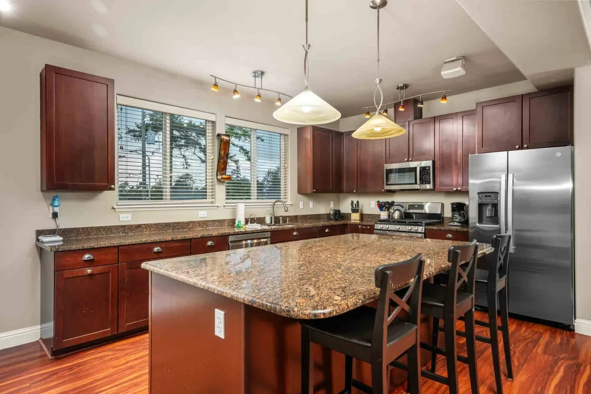 A bright, modern kitchen area showcasing a granite countertop island, stainless steel appliances, and wooden cabinets. The space feels welcoming and practical for meal preparation, baking, or hosting, while maintaining a connection with the larger great room.