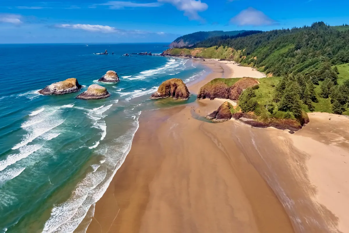 A sweeping view of Seaside’s sandy beach bordered by rocky formations and lush greenery, with waves crashing gently onto the shore. The expansive sand provides plenty of space for building sandcastles, flying kites, or simply relaxing under the sun.