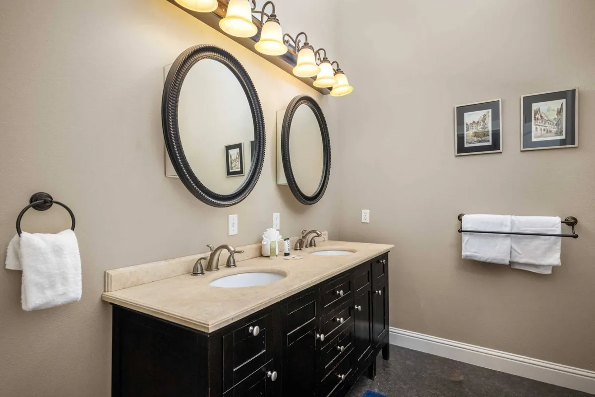 A modern bathroom featuring a double vanity with two sinks, large round mirrors, stylish light fixtures, and tasteful framed artwork. The space is bright, fresh, and evokes a spa-like atmosphere.